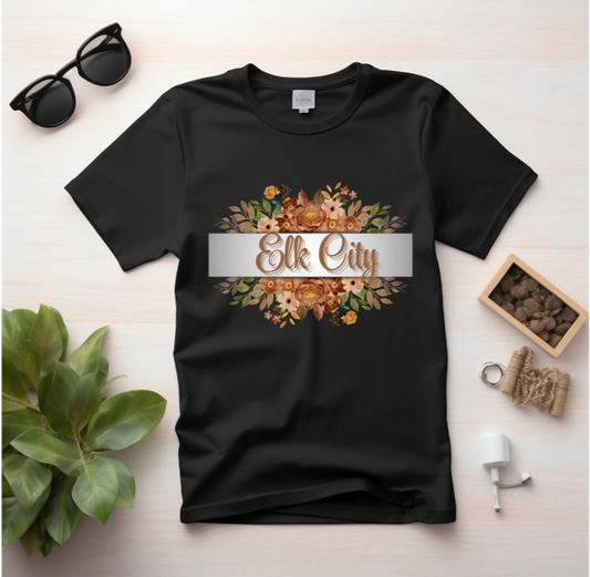 Black t-shirt with 'Elk City' design on a white surface with sunglasses and a plant.